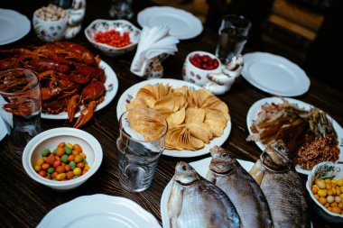 Haşlanmış kerevit, tuzlanmış balık, füme balık, cips, fıstık, ahşap bir masada beyaz tabakta atıştırmalıklar.