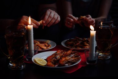 İki bardak bira, iki mum, karides ve limonlu ıstakoz, ellerinde karides.