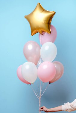 Beyaz, pembe, şeffaf helyum balonları ve altın yıldızın karışımı mavi arka plandaki bir kadının elinde..