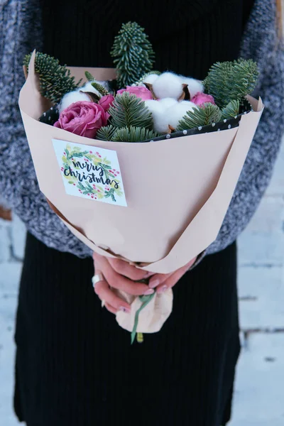 Çiçek buketi, köknar dalları ve şeftali paketinde pamuk kutuları. Arka planda örülü süveter giyen çiçekçi kadının ellerinde. Beyaz tuğla duvar..