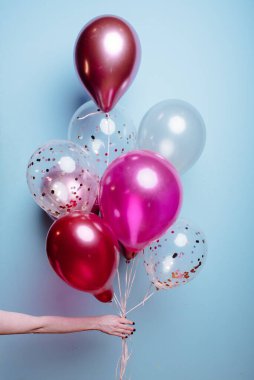 Mavi arka plandaki bir kadının elinde beyaz, pembe, şeffaf helyum balonları var..