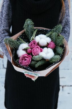 Çiçek buketi, köknar dalları ve şeftali paketinde pamuk kutuları. Arka planda örülü süveter giyen çiçekçi kadının ellerinde. Beyaz tuğla duvar..