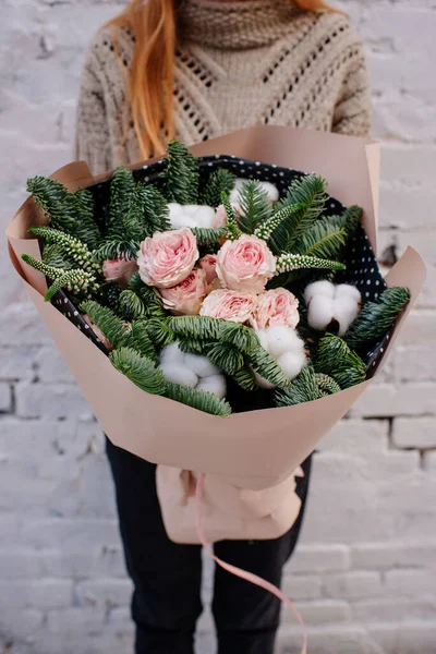 Çiçek buketi, köknar dalları ve şeftali paketinde pamuk kutuları. Arka planda örülü süveter giyen çiçekçi kadının ellerinde. Beyaz tuğla duvar..