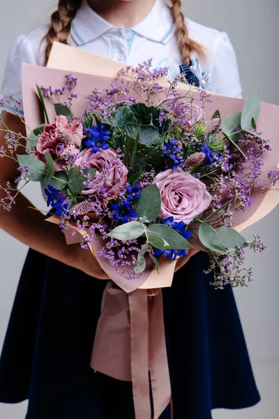 Mor kağıda sarılı bir buket çiçek, açık arkaplanda örgülü saçları olan bir kızın ellerinde. Gül buketi, okaliptüs, karanfil, limonyum, Brassica