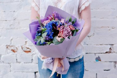 Mor kağıda sarılı bir buket çiçek, arka planda bir kadının ellerinde. Okaliptüs buketi, karanfil, ortanca, gül, Clematis, Cotinus, Gladiolus