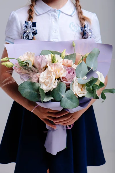 A bouquet of flowers, packed in purple paper in the hands of a girl with pigtails on a light background . Bouquet of Rose, Eucalyptus, Eustoma, Stipa