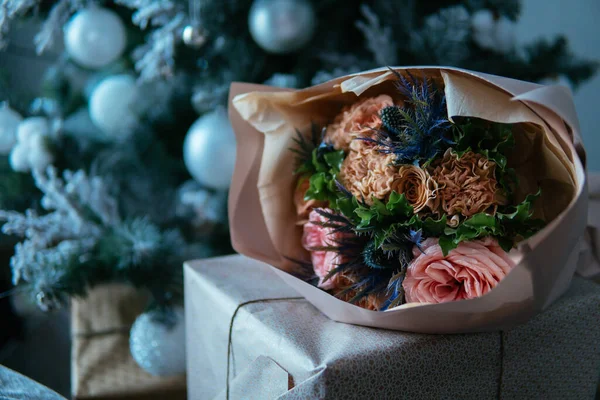 Noel ağacının arka planında el işi kağıda sarılmış şeftali paketinde bir buket çiçek. Rosa, karanfil, eryngium, okaliptüs.