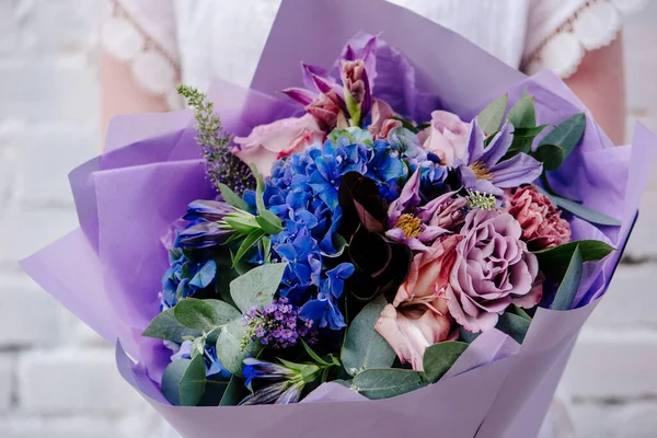 Mor kağıda sarılı bir buket çiçek, arka planda bir kadının ellerinde. Okaliptüs buketi, karanfil, ortanca, gül, Clematis, Cotinus, Gladiolus
