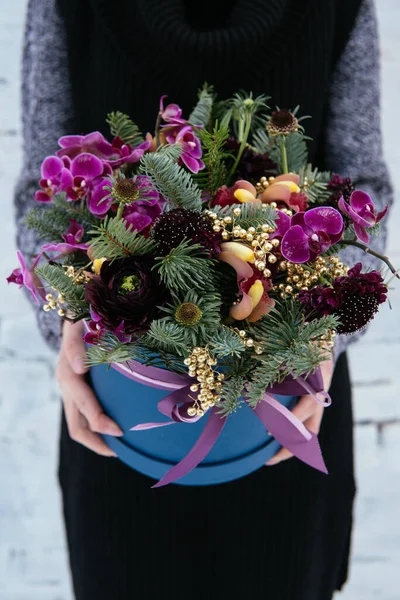 Çiçek buketi ve köknar dalları mavi şapka kutusunda arka planda örülü süveter giyen çiçekçi kadının ellerinde beyaz tuğla duvar..
