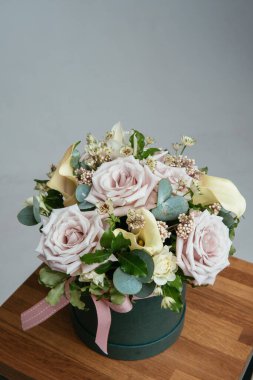 Bir buket çiçek yeşil şapka kutusunda, arka planda tahta koltukta. Rosa, calla, astrantia, okaliptüs, ozotamnus.