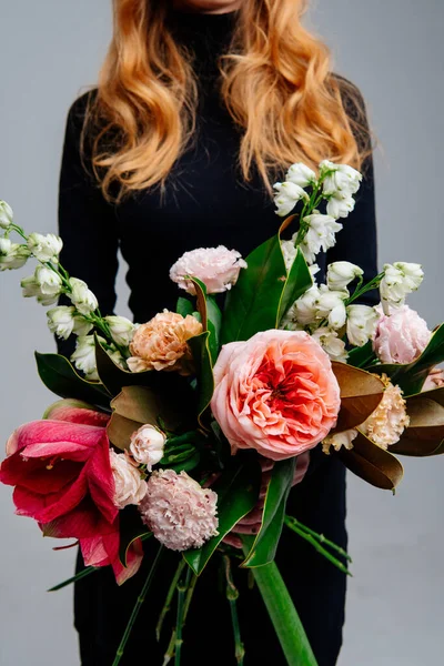 Açık arkaplandaki kızıl saçlı çiçekçinin ellerinde bir buket çiçek. Gül, amilis, östoma, delinyum, manolya yaprakları..