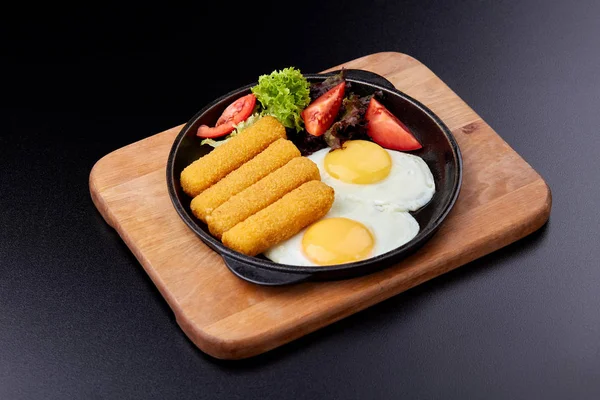 Fried fish sticks or breaded fish fillet in a cast-iron pan, close-up. Fried eggs, fresh tomatoes and salad.