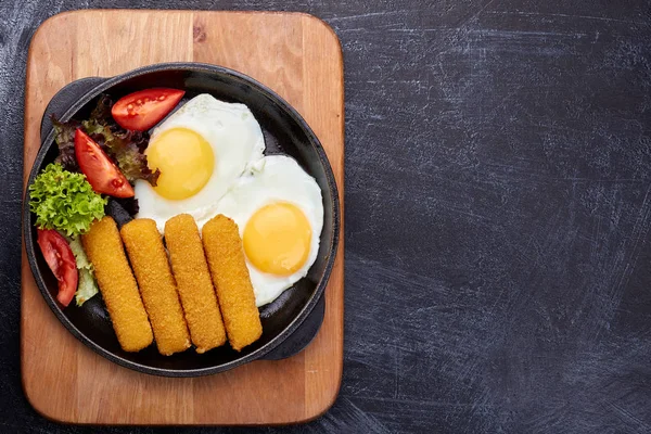 Fried fish sticks or breaded fish fillet in a cast-iron pan, close-up. Fried eggs, fresh tomatoes and salad.