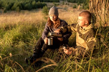 Genç bir adam ve kız arkadaşı parkta termostan çay içiyorlar. Ağacın altında oturup gün batımını izliyorlar. Dinleniyorlar ve rahatlıyorlar..