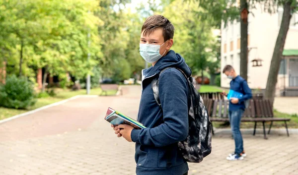 Un adolescente con una máscara médica con libros en las manos en el patio de la escuela. —  Fotos de Stock