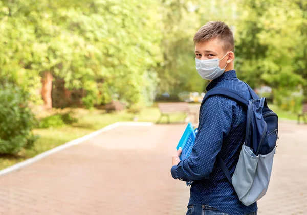 Un adolescente con una máscara médica con libros en las manos en el patio de la escuela. — Foto de Stock