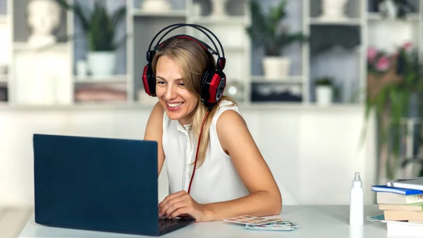 Güzel sarışın bir kız masasında oturmuş kulaklık takıyor ve laptopunu kullanıyor..