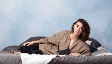 Güzel bir esmer kadın elinde akıllı bir telefonla yatak odasında uzanıyor..