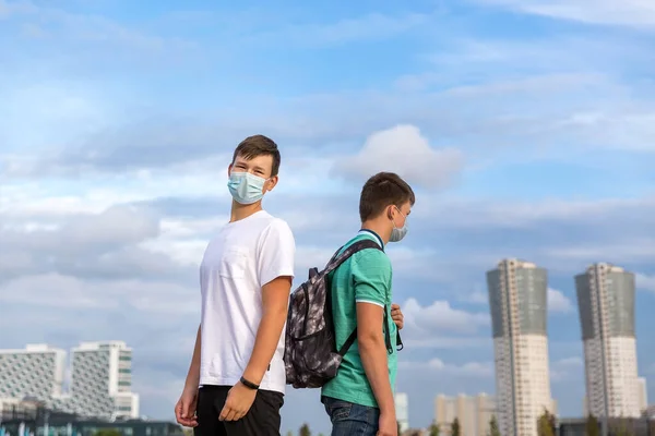 İki arkadaş şehirde yürüyorlar. Yüzlerinde tıbbi maskeler var. Çocuklar doğrudan kameraya bakıyor.. — Stok fotoğraf