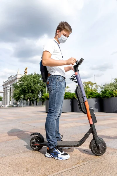 Bir genç parkta scooter 'a biniyor. Yüzü tıbbi bir maskenin içinde. Dikey fotoğraf.