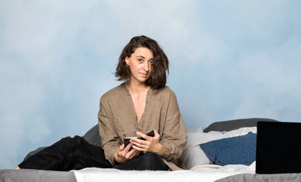Una Hermosa Morena Está Tumbada Cama Dormitorio Con Smartphone Las — Foto de Stock