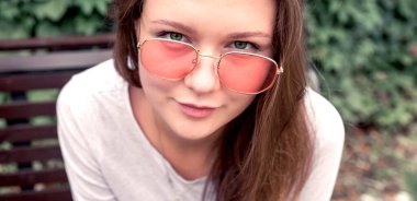 Yeşil bitkilerin arka planında Sunny Pembe gözlüklü güzel kız. Kız kameraya bakıyor. Yakından çekilmiş..