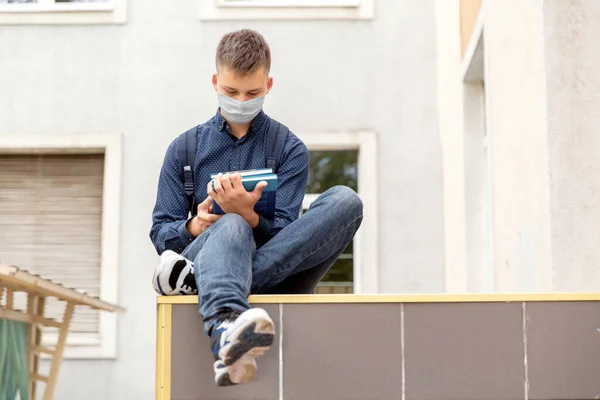 Teen boy sitting in the background of the school. He reads textbooks. His face in a medinah face mask. — Stock Photo, Image