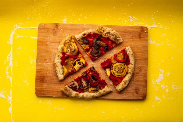 A Rustic and Homemade Vegan Pizza, Two Pieces with Mushrooms and other two with Zucchini and Bell Peppers on a Cutting Board on a Yellow Background