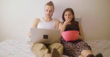 Beyaz bir adam ve İspanyol bir kadın dizüstü bilgisayardan film izliyorlar ve Pembe bir kasede patlamış mısır yiyorlar.