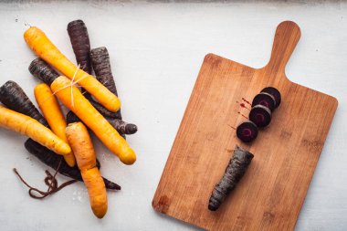 Beyaz arkaplanda havuç parçaları olan kesme tahtasının yanında mor ve sarı havuçlar.