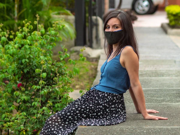 身穿深色手工面罩的拉美裔年轻女性正坐在人行道上 看着摄像机 — 图库照片