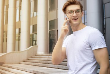 Gözlüklü ve beyaz tişörtlü genç bir öğrenci sokakta oturuyor ve telefonda konuşuyor.