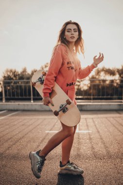 Kırmızı kapüşonlu kaykaylı ve spor ayakkabılı şehir modasına uygun genç bir kız yazın gün batımında yol kenarında park yerinde yürüyor. Aktif spor kadın modelinin açık hava portresi
