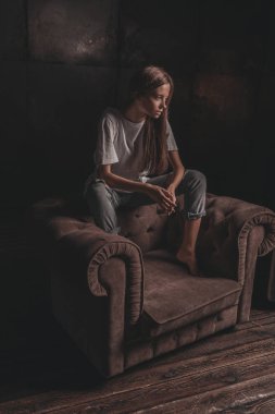 Gömlekli ve kot pantolonlu seksi bir kadın siyah arka plandaki koltukta çatı katında poz veriyor. Baştan çıkarıcı cazibeli genç kız modeli portresi.