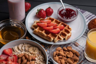 Kahvaltıda. Sağlıklı yiyecek - Belçika waffle 'lı sütlü yulaf ezmesi, meyve, bal, çilek reçeli, çikolatalı kek ve taze yoğurt. Yemeği siyah masa arkasına koyun. Üst görünüm
