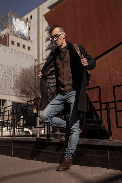 Man street style. Casual clothes. Young student wearing in coat, jeans, boots,backpack, glasses.