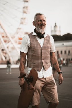 Kaykaylı şık hippi sakallı adam. Günlük kıyafetlerle yetişkin modeli şehir caddesinde uzun tahta sürüyor. Sokak yaşamı spor fotoğrafı.