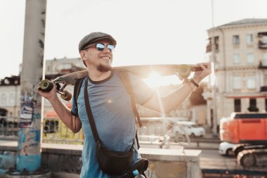 Günbatımında şehir sokağında poz veren kaykaylı şık yakışıklı bir adam. Gülümseyen modern erkek portresi. Kaykaycı şehir yaşam tarzını seviyor