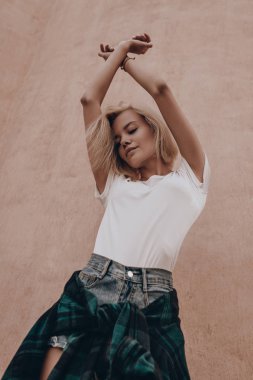 Beyaz tişörtlü, mavi şortlu seksi sarışın kız şehir caddesinde duvarın yanında poz veriyor. Şehirli arka planda yürüyen baştan çıkarıcı, yakışıklı güzel kadın.