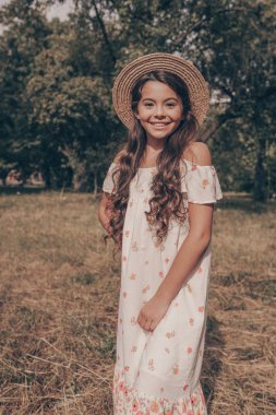 Genç güzel kız parkta yürüyor. Şapka portresindeki şık bir bebek. Gülümseyen bir çocuk yazın yeşil ağaçların arkasında dans ediyor.