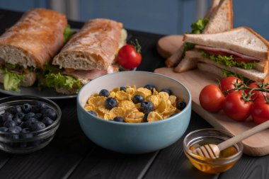 Breakfast set. Fresh meal. Food. Traditional sandwich, delicious baguette and corn flakes with milk, fruit and honey on black table. Kitchen
