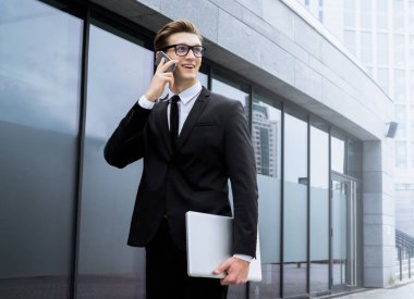 Genç ve yakışıklı bir iş adamı telefonunu kullanırken dizüstü bilgisayarını tutuyor ve sokakta dikiliyor.