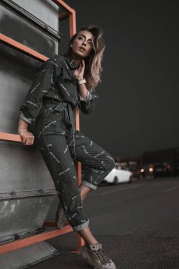 Kot pantolon ve spor ayakkabılarıyla neon ışıkta park yerinde poz veren şık bir kızın moda sokak portresi. Geceleri park yerinde poz veren modern genç kadın modeller. Kentsel yaşam tarzı