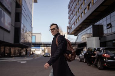 Şehir caddesinde yürüyen genç ve şık bir adam. Kentsel tarzı fotoğraf çekimi. Siyah palto, gözlük ve sırt çantası giymek.