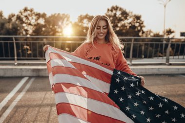 Kırmızı kapüşonlu, şehir modasına uygun güzel bir kız Amerikan bayrağıyla park yerinin yakınında yürüyor. Mutlu kadın güzellik portresi yaz günbatımında