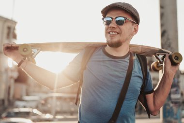 Sakallı hippi adamın portresi mavi gömlekli, gri şapkalı ve güneş gözlüklü şehir caddesinde. Arkaplanda yol kenarında poz veren mutlu model.