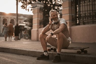 Yakışıklı yetişkin bir adam uzun panoda oturuyor ve şehir caddesinde günlük kıyafetlerle telefonla konuşuyor. Açık havada kaykaylı şık bir erkek portresi..