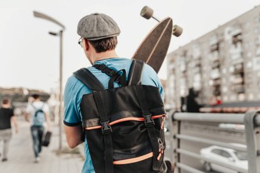 Şehir caddesinde gün batımında poz veren bir patenci. Uzun panolu şık bir hippi binanın arka planında yol kenarında yürüyor. Sakallı spor erkek modelinin şehir tarzı portresi