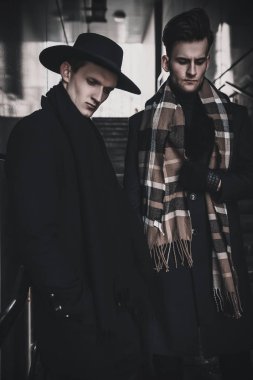İş merkezinin yakınındaki merdivenlerde poz veren iki erkek model. Palto, şapka, eldiven, atkı giyiyor. Erkek model testleri. Sokak stili