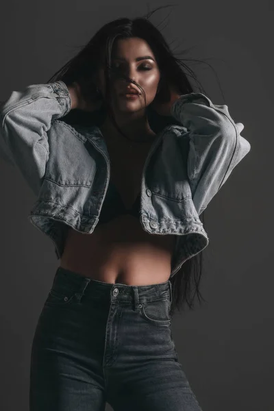 İç çamaşırlı, kot pantolonlu ve kot ceketli seksi bir kadın stüdyoda siyah arka planda poz veriyor. Baştan çıkarıcı çıplak cazibeli genç kız modeli portresi. İç çamaşırı tutkusu.